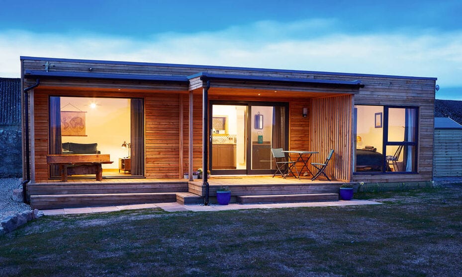 A timber-clad modular building at dusk, with a covered veranda and warmly lit windows